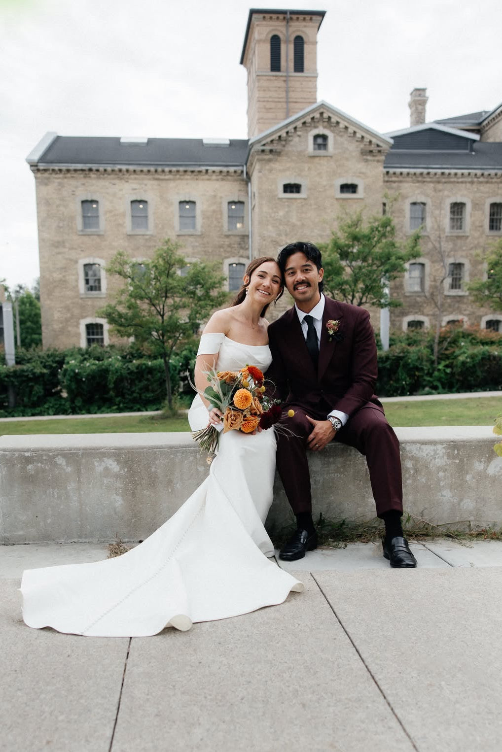 A bride in an off-the-shoulder white wedding gown and a groom in a dark burgundy suit sit together on a concrete ledge outdoors, with a large historic stone building in the background.