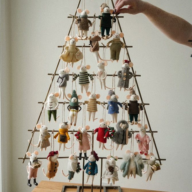 A hand is placing a small hanging ornament onto a rustic metal tree stand decorated with many tiny mouse-like figurines, set on a wooden table against a plain indoor wall.