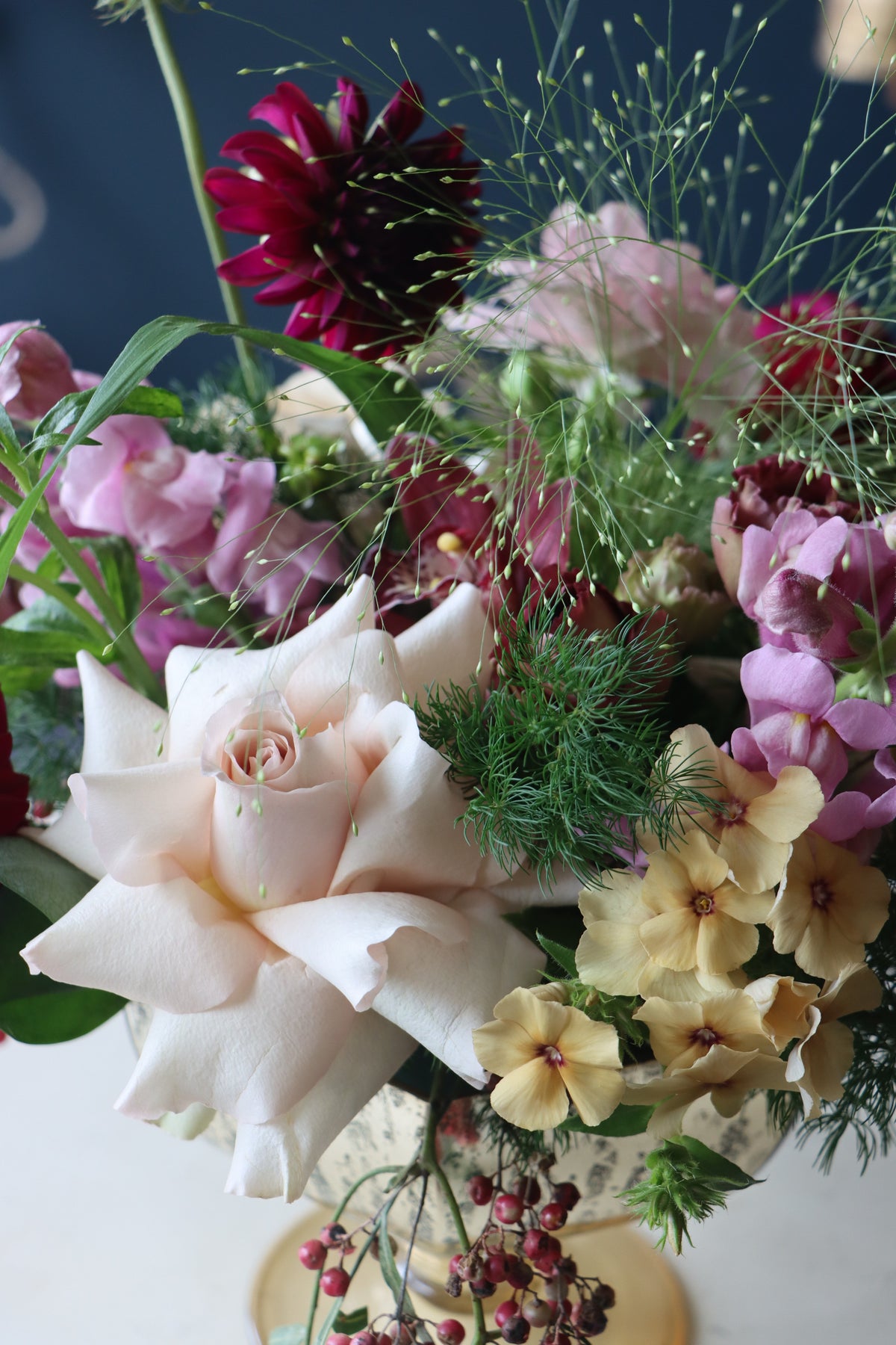A close-up of a mixed floral arrangement featuring a large pale blush rose, deep burgundy dahlias, pink snapdragons, pale yellow flowers, and green foliage.